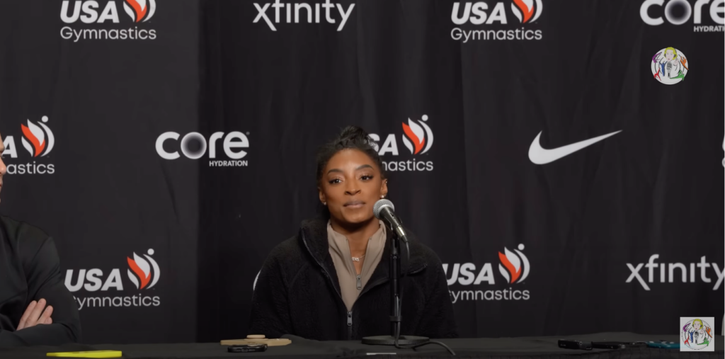 Simone Biles, in a black jacket and tan shirt at a press conference with a USA Gymnastics background. 